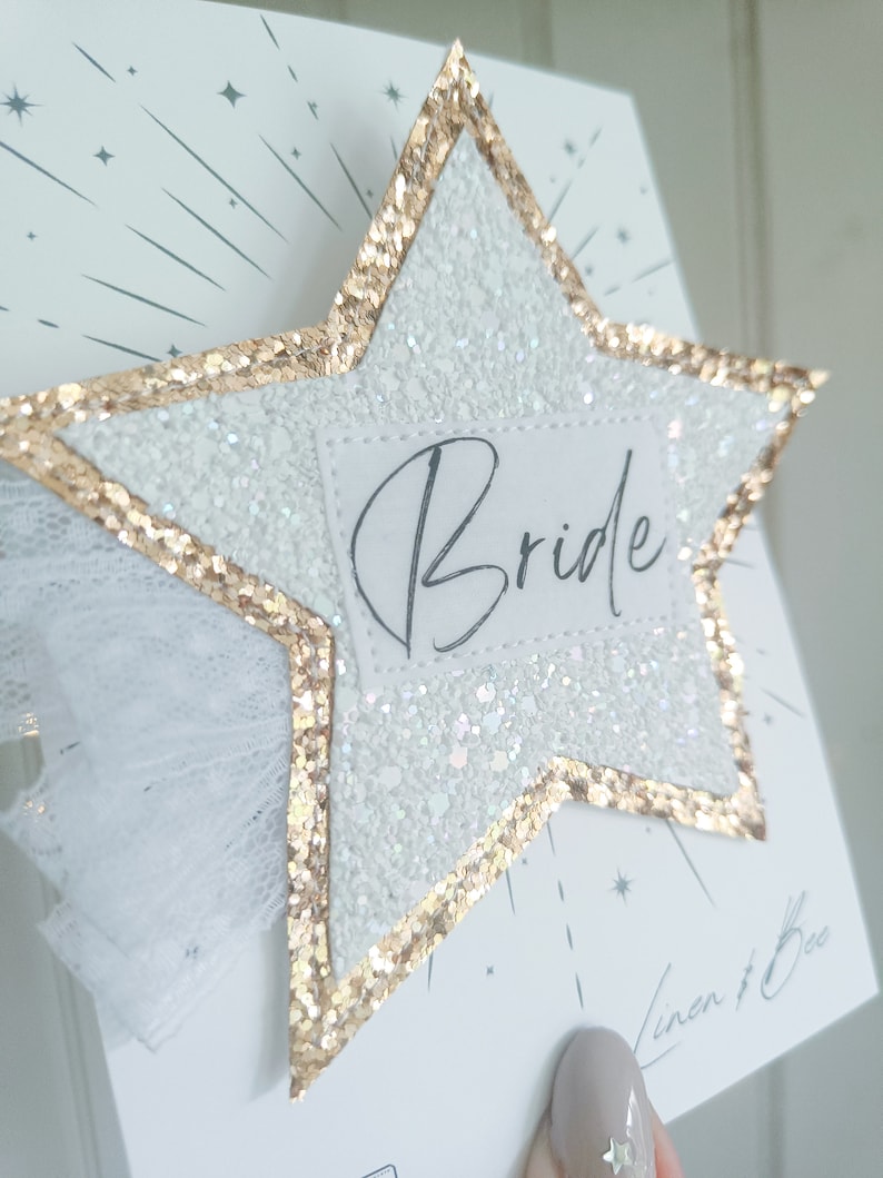 May include: A white glitter star with a gold glitter border and a white tulle bow. The star has a white label with the word "Bride" written in black cursive.