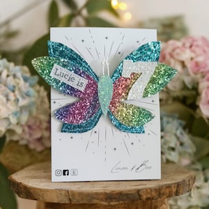 Op de afbeelding: Een regenboog glittervlinder met de tekst "Lucie is 7" erop. De vlinder bevindt zich op een witte kaart met de tekst "Linen & Bee" erop.