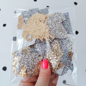 May include: A clear plastic bag filled with a variety of glitter shapes in gold, silver, and iridescent colours. The glitter shapes are in the form of stars, hearts, and other geometric shapes.