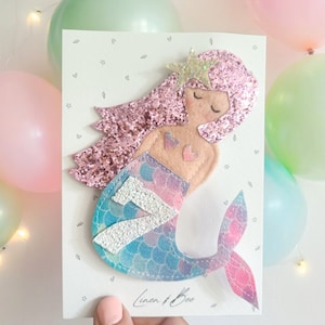 May include: A pink glitter mermaid with a silver glitter number 7 on her tail. The mermaid is on a white card with the text "Linen & Bee".