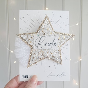 May include: A white and gold glitter star with the word "Bride" written on it. The star has a white tulle bow attached to it. The star is on a white card with the words "Linen & Bee" written on it.