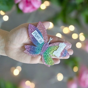 Op de afbeelding: Een regenboog glitter vlinder haarspeld met de tekst "piper is" erop.