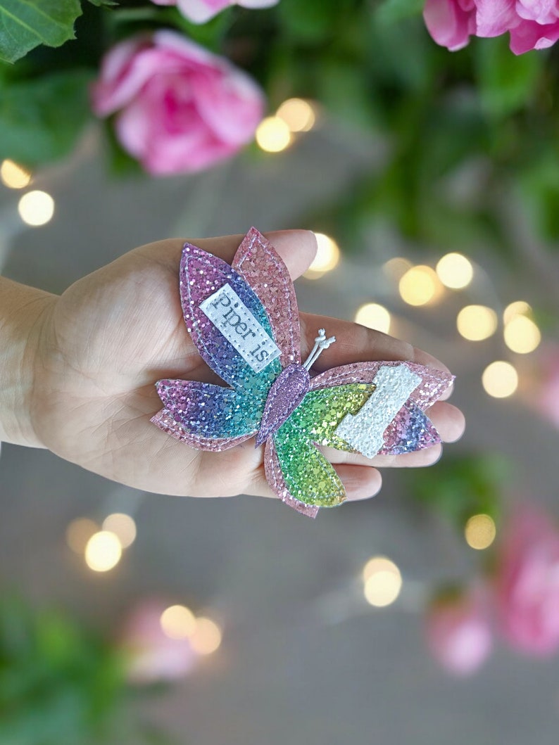 Op de afbeelding: Een regenboog glitter vlinder haarspeld met de tekst "piper is" erop.