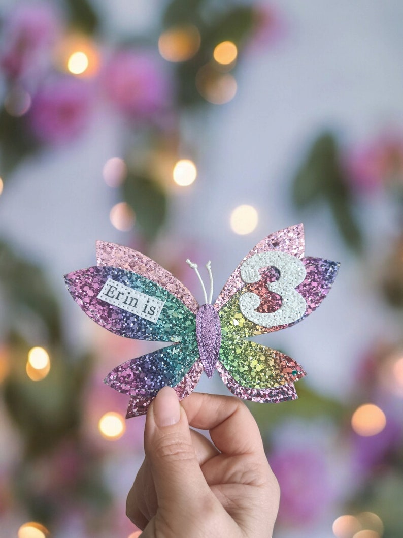 Op de afbeelding: Een regenboog glittervlinder met de tekst "Erin is 3" op een vleugel. De vlinder wordt in een hand gehouden tegen een wazige achtergrond van roze en groen.