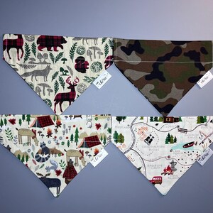 May include: Four different patterned dog bandanas with a white background. The bandanas feature a variety of designs, including a red and black plaid pattern with deer, a camouflage pattern, a camping scene with tents and trees, and a map of a campground with trails and landmarks. The bandanas are all trimmed with white stitching and have a small tag with the word "cherie" on them.