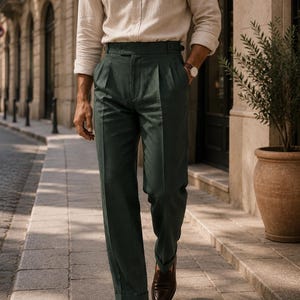 May include: A beige long-sleeved button-down shirt with a collar and dark forest green trousers featuring a high waist, double pleats, and cuffed hems. The trousers have side adjusters at the waistband and are paired with dark brown leather loafers.