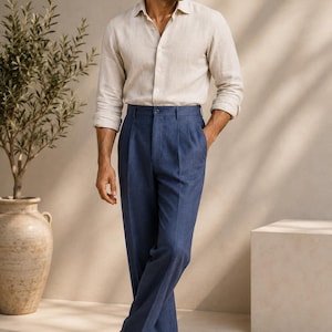 May include: A beige long-sleeved button-down shirt paired with high-waisted blue trousers featuring front pleats and a straight-leg cut. The outfit includes black leather loafers. A logo for Atelier Suits Co. is visible in the bottom corner.