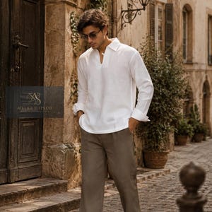 May include: A long-sleeved white shirt with a collar and V-neck opening paired with loose-fitting olive green trousers and white trainers. A logo with the text ATELIER SUITS CO. is visible on the left side of the image over the clothing.