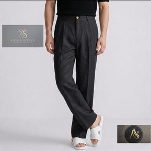 May include: Charcoal gray trousers featuring a high-waisted design with front pleats, a single button closure, and a straight-leg cut. The garment includes a logo patch with the text ATELIER SUITS CO. and a circular emblem with the letters AS.
