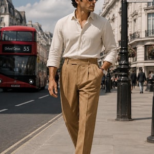 May include: A long-sleeved off-white button-down shirt with a collar and tan high-waisted trousers featuring pleats at the waist. The outfit includes dark brown leather loafers and black sunglasses, worn with a wristwatch on the left wrist.
