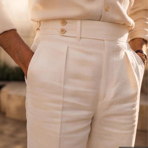 May include: Off-white trousers featuring a high-waisted design with a wide waistband, double button side adjusters, and front pleats. The trousers are paired with a matching off-white long-sleeved shirt with a single visible button at the waist.