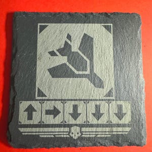 May include: A square, dark gray slate coaster with a light gray graphic design. The design includes a stylized symbol, directional arrows, and a skull emblem. The coaster is set against a red background.