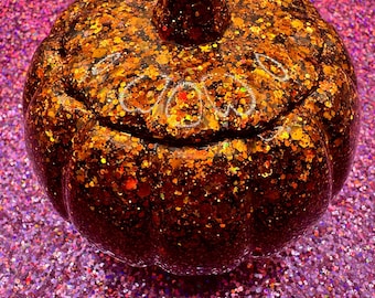 May include: A glitter-covered pumpkin-shaped container with a lid. The pumpkin is primarily orange and brown with flecks of gold and red glitter. The background is a sparkly purple surface.