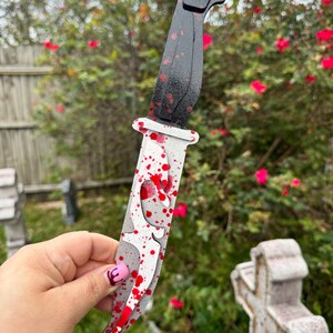 May include: A decorative knife with a black handle and a silver blade, splattered with red paint. The blade has a unique cutout design. The knife is held in front of a blurred background of green foliage and red flowers.