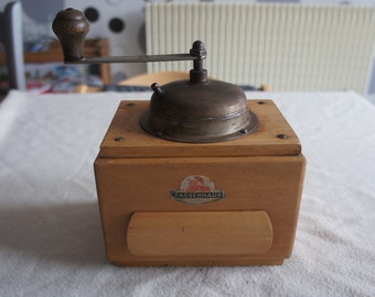Könnte beinhalten: Eine antike Kaffeemühle aus Holz mit einem Metallmahlwerk und einem Kurbelgriff. Die Mühle hat eine Schublade und ein Etikett mit der Aufschrift "ZASSENHAUS".