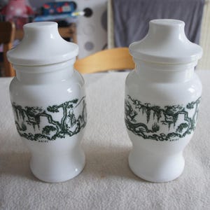 Peut inclure: Deux pots en céramique blancs avec couvercles, ornés d'un motif asiatique vert et noir. Les pots ont une forme cylindrique avec une base évasée et un couvercle arrondi. Le mot "ARIEL" est imprimé sur les pots.