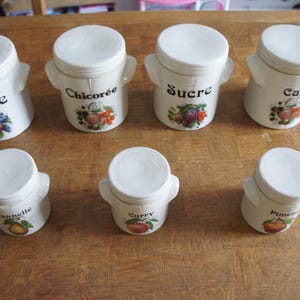 May include: A set of seven white ceramic kitchen canisters with lids and side handles. Each canister features black text and a colourful fruit illustration. The labels include Farine, Chicorée, Sucre, Café, Cannelle, Curry, and Piment.