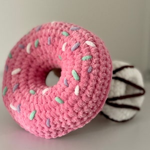 Puede incluir: Una almohada de donut rosa hecha a crochet con chispas de colores. El donut está sobre una superficie blanca.