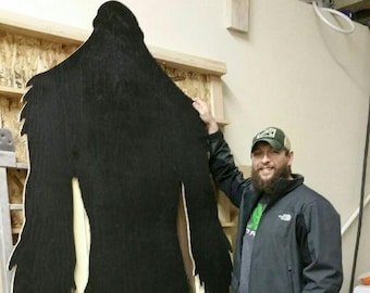 May include: A black silhouette of Bigfoot, a mythical creature, is mounted on a wooden frame. A person wearing a black jacket, blue jeans, and a green and black baseball cap stands next to the silhouette.