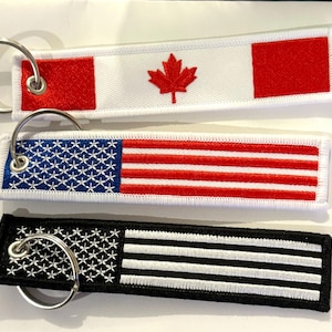 May include: Three keychains featuring different flag designs. The top keychain has a Canadian flag with a red maple leaf on a white background. The middle keychain has an American flag with red and white stripes and blue stars. The bottom keychain has a black and white striped flag.