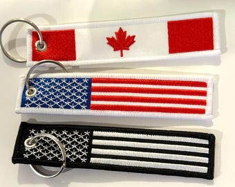 May include: Three keychains with different flag designs. The top keychain features a Canadian flag with a red maple leaf on a white background. The middle keychain features an American flag with red and white stripes and blue stars. The bottom keychain features a black and white striped flag.