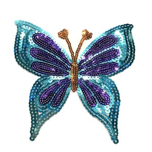 May include: A sequined butterfly applique in shades of blue and purple. The wings are outlined with light blue sequins, with purple detailing. The body and antennae are gold-coloured. Ideal for adding a decorative touch to garments or crafts.