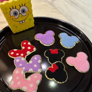 May include: Eight felt Mickey Mouse-themed patches in various colors, including pink, purple, red, black, and blue. The patches have gold glitter trim and some have white polka dots.