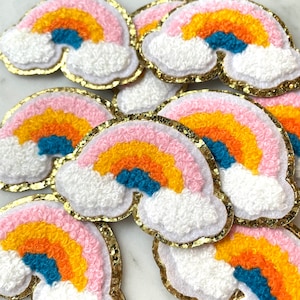May include: A collection of rainbow-coloured felt patches with a fluffy white cloud design. Each patch has a gold glitter trim.