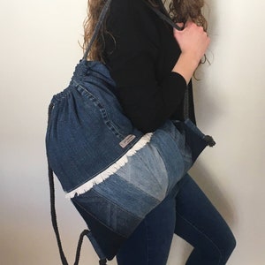 Puede incluir: Una mochila de nailon con cordón de ajuste y ribete de flecos blancos. La mochila está hecha de nailon reciclado y tiene un diseño único de patchwork.