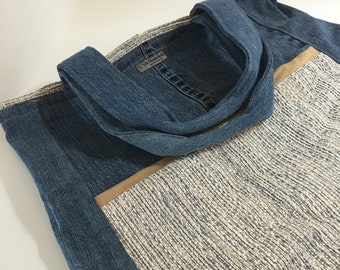 May include: A denim tote bag with a woven fabric panel. The bag features a blue denim exterior with a woven fabric panel in shades of beige and grey. The handles are made of denim. The bag is designed for carrying items.