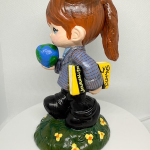 May include: A ceramic figurine of a child holding a globe and books labeled "Journalism" and "for Dummies." The child wears a blue plaid jacket, black boots, and stands on a grassy base with yellow flowers.