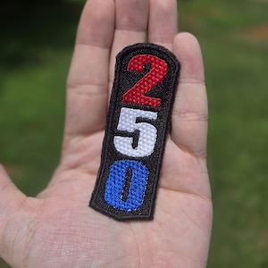 Puede incluir: Un parche bordado rectangular con fondo negro que muestra el número 250 apilado verticalmente. El número 2 está bordado en rojo, el 5 en blanco y el 0 en azul, todo ello rodeado por un borde negro grueso en los bordes del parche.