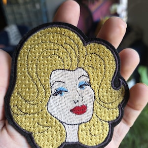 Puede incluir: Un parche bordado que presenta el retrato estilizado de una persona con voluminoso cabello amarillo dorado, sombra de ojos azul brillante, largas pestañas oscuras y labios rojos intensos, todo delineado con un borde negro grueso sobre tela blanca.