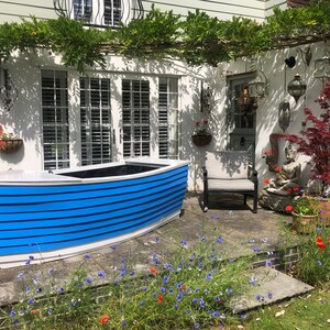 May include: A blue and white wooden boat planter with a black liner. The planter is sitting on a patio with a stone floor and surrounded by flowers.