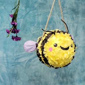 May include: A yellow and black bee-shaped pinata with a smiling face and purple wings hanging from a string.