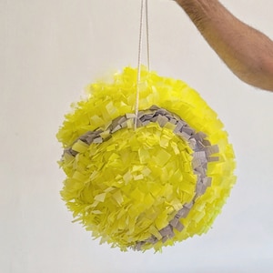 May include: A yellow and grey tennis ball shaped pinata hanging from a white string.