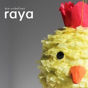 Könnte beinhalten: Eine gelbe Küken-Piñata aus Fransenpapier mit einer roten Papierkrone auf dem Kopf, zwei schwarzen runden Augen und einem orangefarbenen Papierschnabel. Der Text "éco-créations raya" ist oben links vor einem grauen Hintergrund zu sehen.