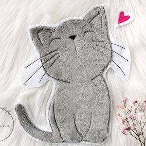 Könnte beinhalten: Ein grauer Plüschkatze mit einem weißen Umriss und einem rosa Herz darüber. Die Katze hat ein lächelndes Gesicht und geschlossene Augen.