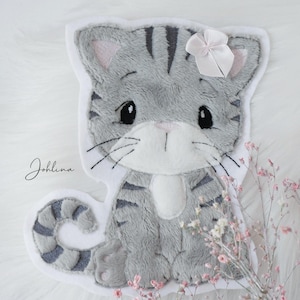 Könnte beinhalten: Ein kissenförmiges graues Kätzchen mit schwarzen Streifen und einer weißen Schleife. Die Katze hat rosa Innenohren und schwarze Schnurrhaare. Das Wort "Johlina" ist links in Kursivschrift geschrieben. Rosa Blumen sind im Hintergrund.