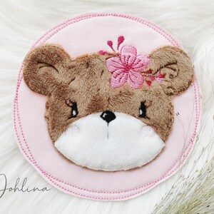 Könnte beinhalten: Ein kreisförmiger Stoffaufnäher mit einem gestickten braunen Teddybärenkopf, weißer Schnauze, schwarzen Augen und einer rosa Blume am Kopf. Der Bär ist auf einem hellrosa Hintergrund mit genähtem Rand zentriert. Der Text Johlina ist sichtbar.