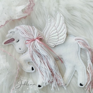 Könnte beinhalten: Weißes Filz-Einhorn mit rosafarbenem Glitzerhorn und rosafarbenem Garnmähne und -schwanz. Das Einhorn hat weiße Filzflügel mit rosa Nähten.