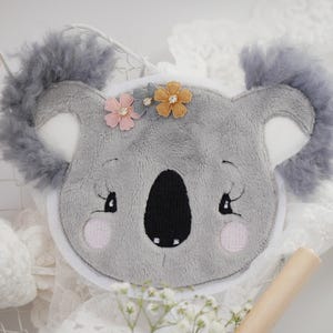 Könnte beinhalten: Ein graues Koala-Gesicht aus Plüsch mit flauschigen Ohren und gestickten Details. Der Koala hat eine schwarze Nase, rosa Wangen und ist mit kleinen rosa und gelben Blumen verziert. Wahrscheinlich ein dekoratives Kissen oder eine Applikation.