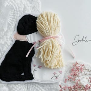 Könnte beinhalten: Ein schwarzer Filzhund mit einem weißen Filzkleid und blonden Garnhaaren umarmt sich. Die Filzfiguren sind auf einen weißen Stoffhintergrund mit rosa Nähten genäht. Der Text "Johllina" ist in schwarzer Tinte auf den weißen Stoff geschrieben.