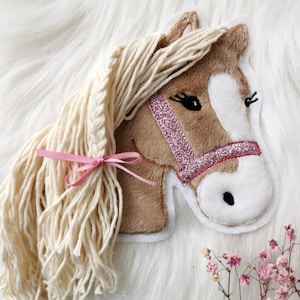 Könnte beinhalten: Ein Plüschpferdekopf mit cremefarbener Mähne und einem rosa Glitzerhalfter. Das Pferd hat eine weiße Schnauze und schwarze Augen. Ein rosa Band ist um die Mähne gebunden. Ein dekorativer Artikel.