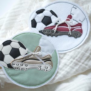 Könnte beinhalten: Zwei kreisförmige Stoffaufnäher mit gestickten Fußballmotiven. Ein Aufnäher hat einen weißen Hintergrund mit einem schwarz-weißen Fußball und einem kastanienbraunen Fußballschuh. Der andere hat einen salbeigrünen Hintergrund mit Ball und beigem Schuh.
