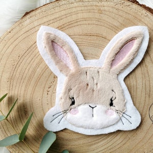 Könnte beinhalten: Ein beigefarbener Plüsch-Hasen-Applik mit gestickten Augen und Schnurrhaaren. Der Hase hat rosa Ohren und ein weißes Gesicht.
