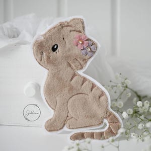 Könnte beinhalten: Ein Applikation in Form einer Katze aus beigem Plüsch mit gestickten Details und weißem Rand. Die Katze hat kleine rosa und lila Blumenverzierungen auf dem Kopf. Die Applikation liegt auf einer weißen Oberfläche.