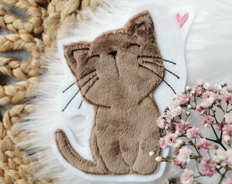 May include: A brown cat-shaped felt applique with black whiskers and a pink heart. The cat is smiling with its eyes closed. The applique is on a white background, with a fluffy white surface and pink flowers in the background.