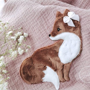 Könnte beinhalten: Ein braunes und weißes Plüschfuchs-Applik mit einer weißen Schleife auf einem rosa Musselin-Stoff. Der Fuchs schaut nach rechts und hat einen weißen Bauch und eine weiße Schwanzspitze.