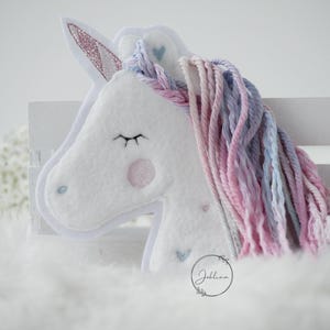 Könnte beinhalten: Weißes Plüschtier in Form eines Einhorns mit einer rosafarbenen und violetten Garnmähne und einem rosafarbenen Glitzerhorn. Das Einhorn hat gestickte Augen und ein rosafarbenes Herz auf der Stirn.